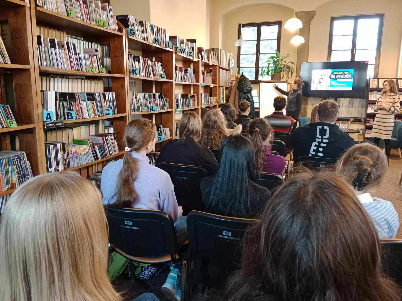 Uczniowie szkół siedzą na krzesłach, słuchają kobiety stojącej przy tablicy. Po bokach, przy ścianach ustawione są regały z książkami.