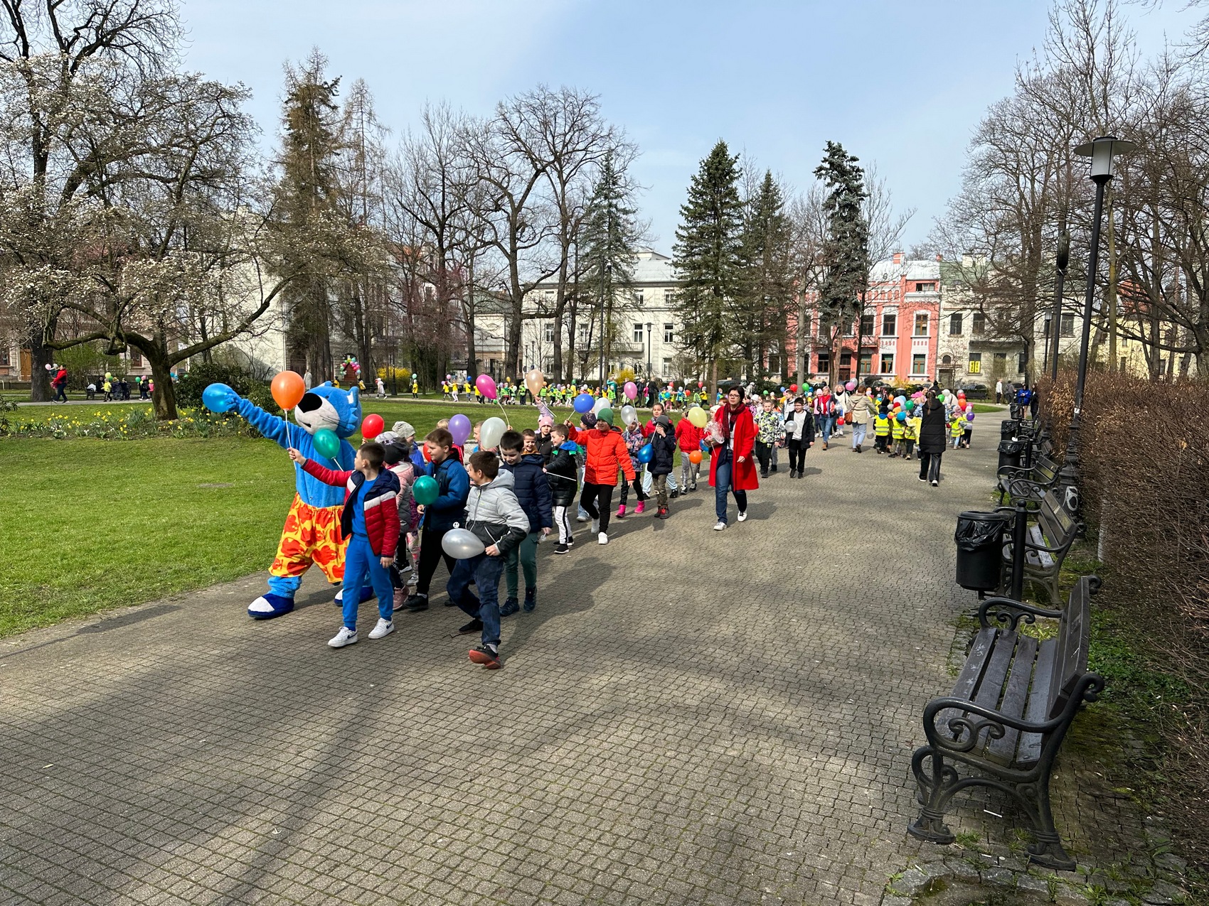 Rozgrzewka czytelnicza_10 Park miejski, duża grupa dzieci maszeruje chodnikiem, trzymając w rękach balony. Całą grupę prowadzi człowiek w przebraniu niebieskiego kota. Na zdjęciu widoczne są kwitnące drzewa, trawy, kwiaty oraz ławki.