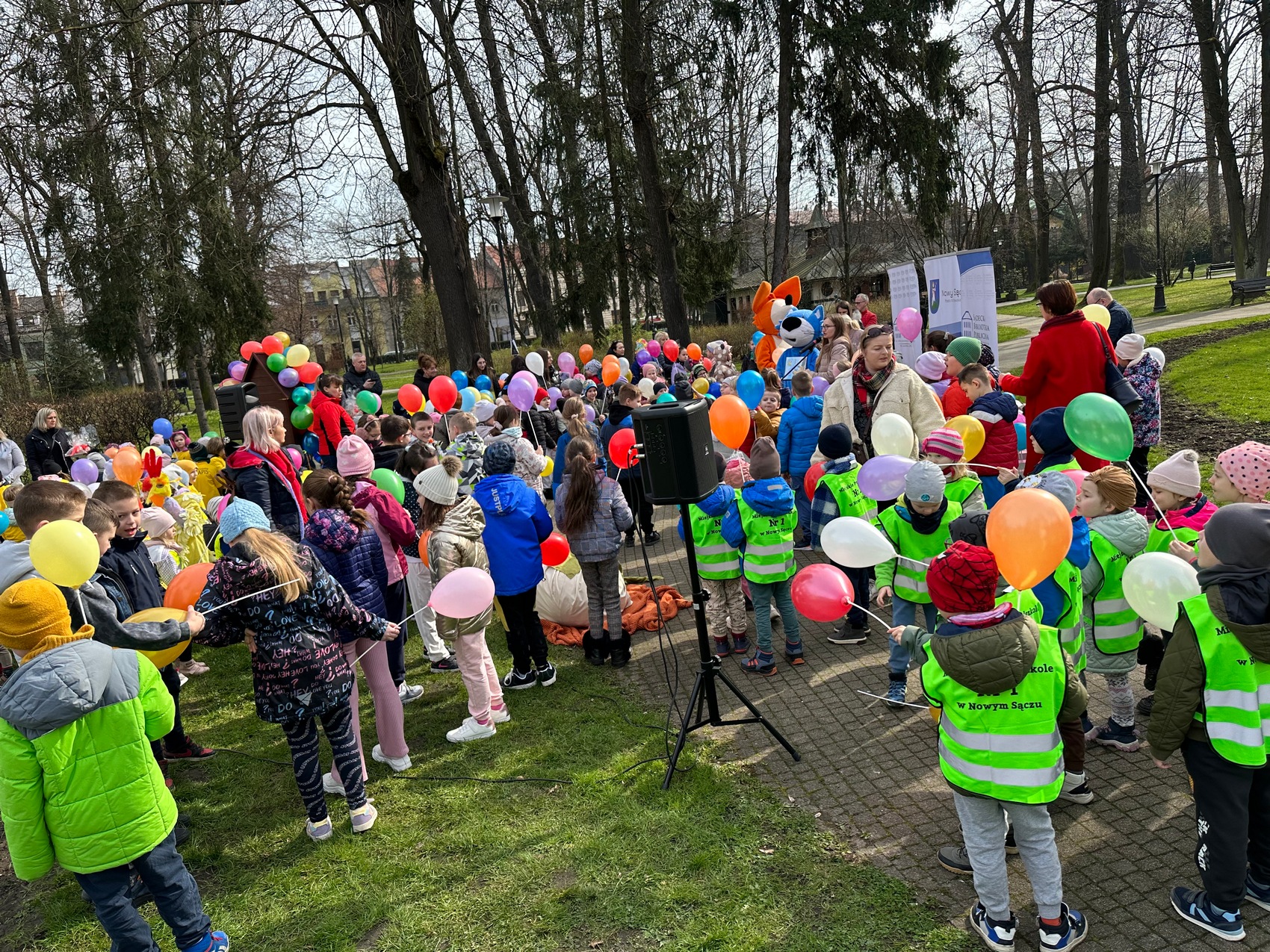 Rozgrzewka czytelnicza_12 Duża grupa dzieci z balonikami w rękach stoi w parku miejskim. Wszyscy ze sobą rozmawiają oraz bawią się balonami. Pomiędzy dziećmi stoją Panie opiekunki, bajkowe postaci: lis oraz kot.