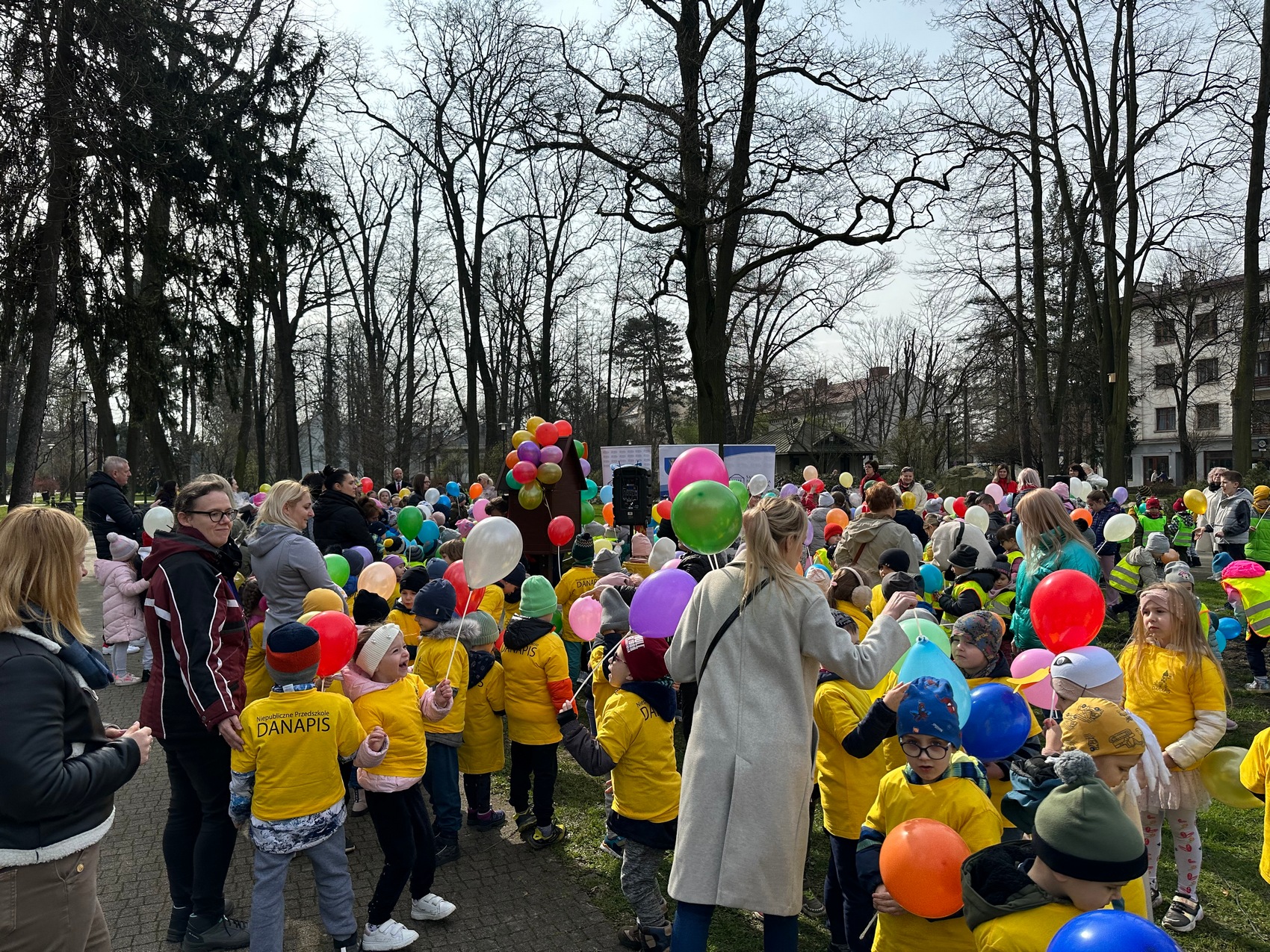 Rozgrzewka czytelnicza_2 Duża grupa dzieci z balonami przechadza się po miejskim parku, wraz z nimi idą Panie opiekunki. W tle widoczne są budynki i drzewa.