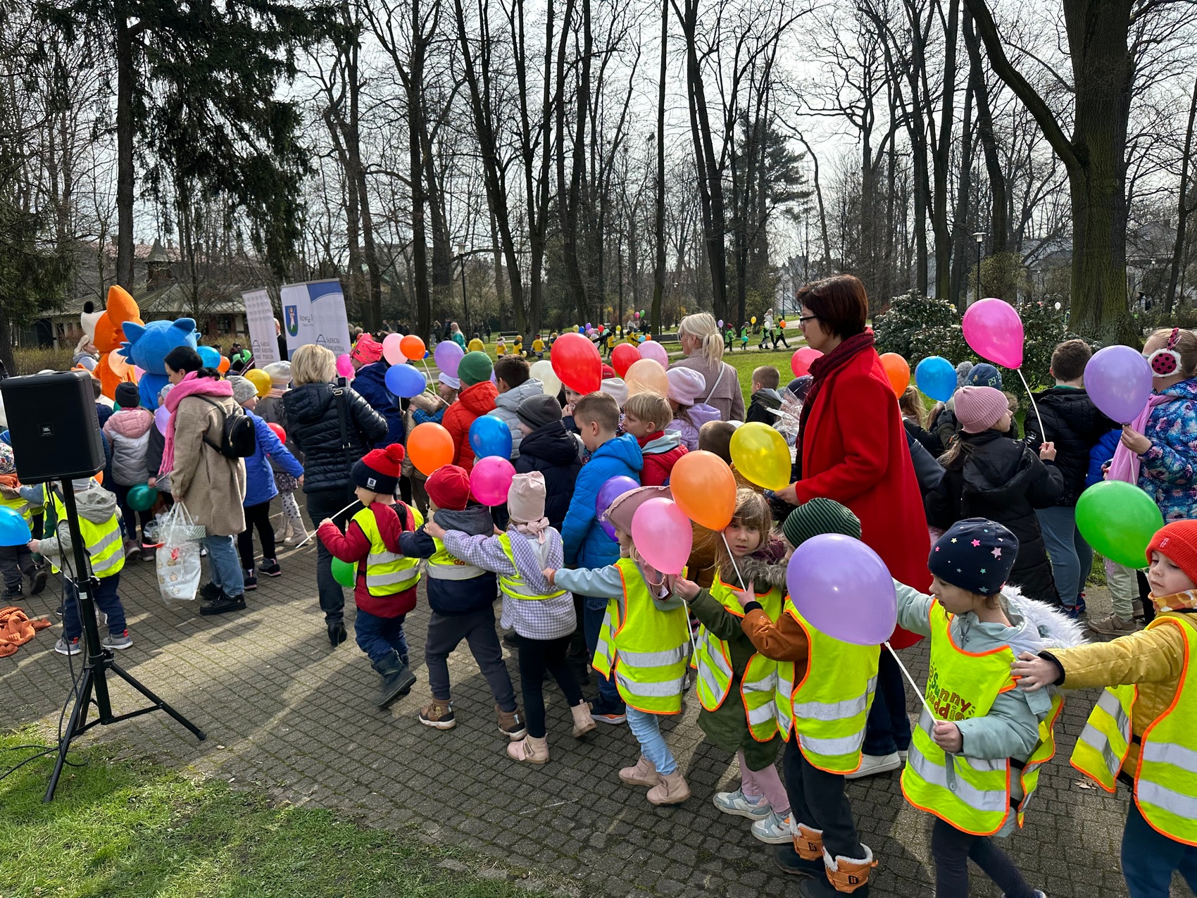 Rozgrzewka czytelnicza_3 Duża grupa dzieci z balonami przechadza się po miejskim parku, wraz z nimi idą Panie opiekunki. W tle widoczne są budynki i drzewa.