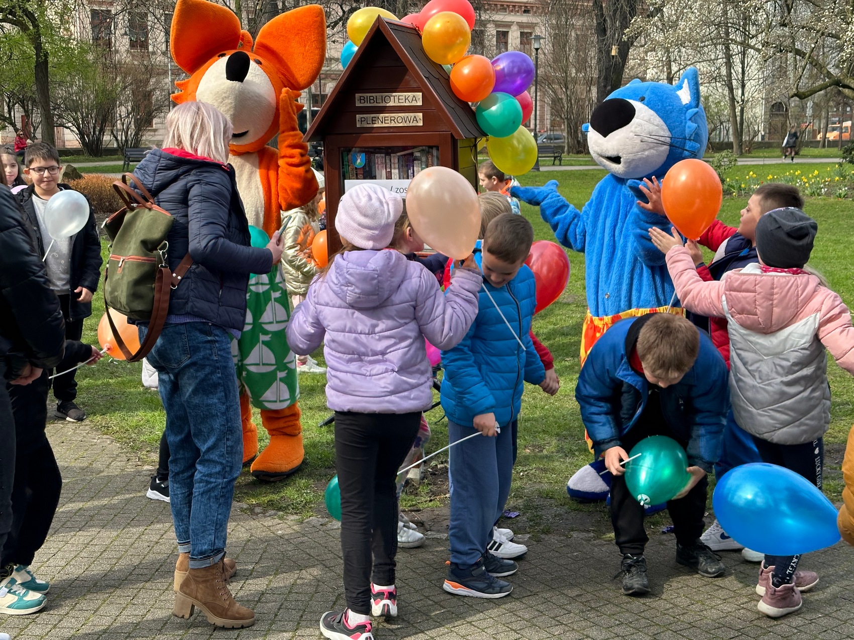 Rozgrzewka czytelnicza_8 Miejski park, bajkowe postaci: lis oraz kot stoją obok domku z książkami, przy nich stoją rozbawione dzieci trzymające balony oraz ich opiekunka.