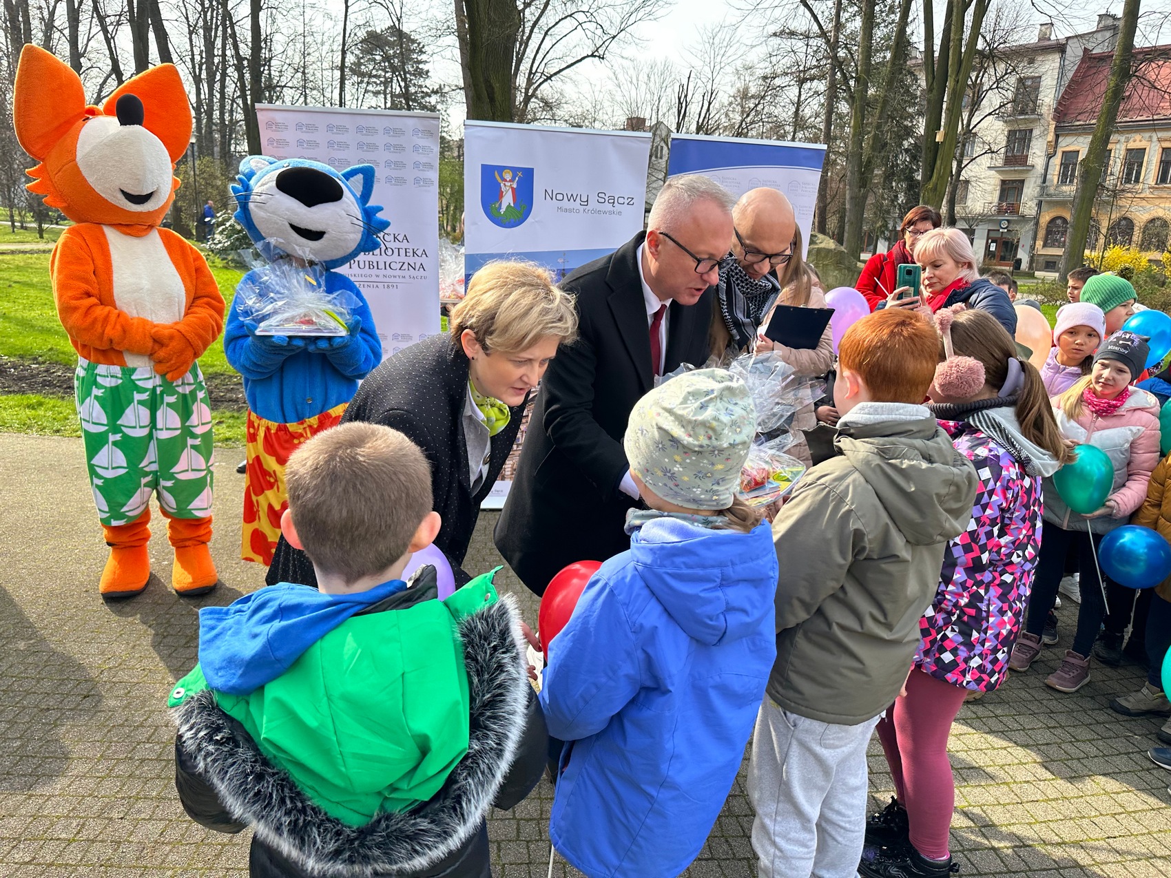Rozgrzewka czytelnicza_9 trójka zaproszonych gości,: kobieta oraz dwóch ,mężczyzn wręczają prezenty dla dzieci. W tle stoją bajkowe postaci: lis oraz kot. Po prawej stronie widoczne są budynki oraz dzieci i kobieta robiąca zdjęcie telefonem.