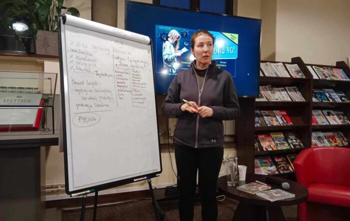 Kobieta stoi przy tablicy z zapiskami. Za nią widnieje ekran, obok stolik z napojem i czerwony fotek. Dalej widoczny jest regał z czasopismami.