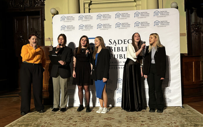 Pięć kobiet śpiewa do mikrofonów. Za nimi widnieje logo Sądeckiej Biblioteki Publicznej.