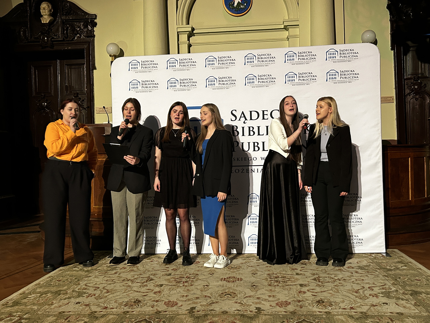 Pięć kobiet śpiewa do mikrofonów. Za nimi widnieje logo Sądeckiej Biblioteki Publicznej.