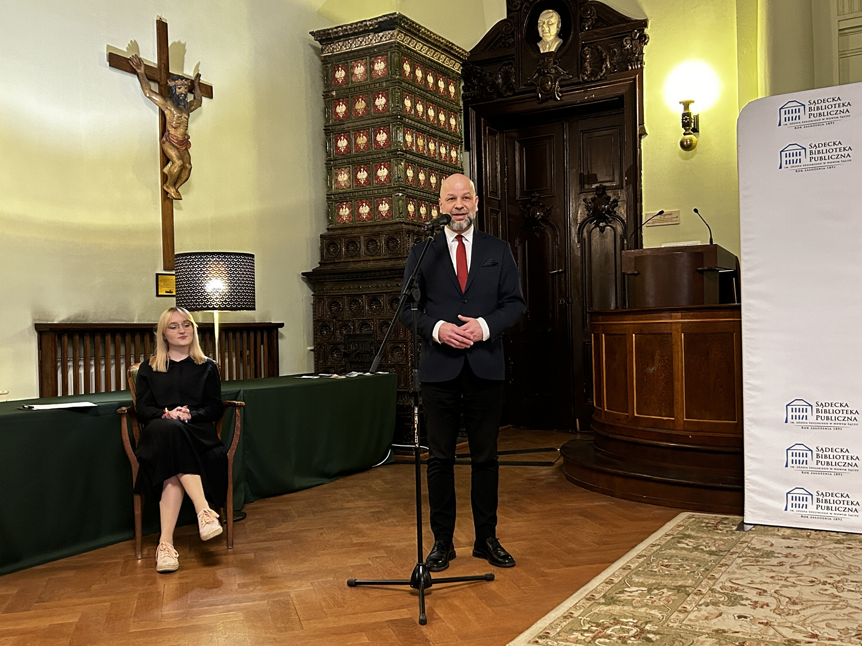 piosenki Jacka Cygana (12) Po lewej stronie siedzi na krześle kobieta o jasnych włosach, po prawej stronie przed mikrofonem stoi mężczyzna. W tle widać zabytkowy komin kaflowy, ogromne, drewniane drzwi oraz fragment mównicy.
