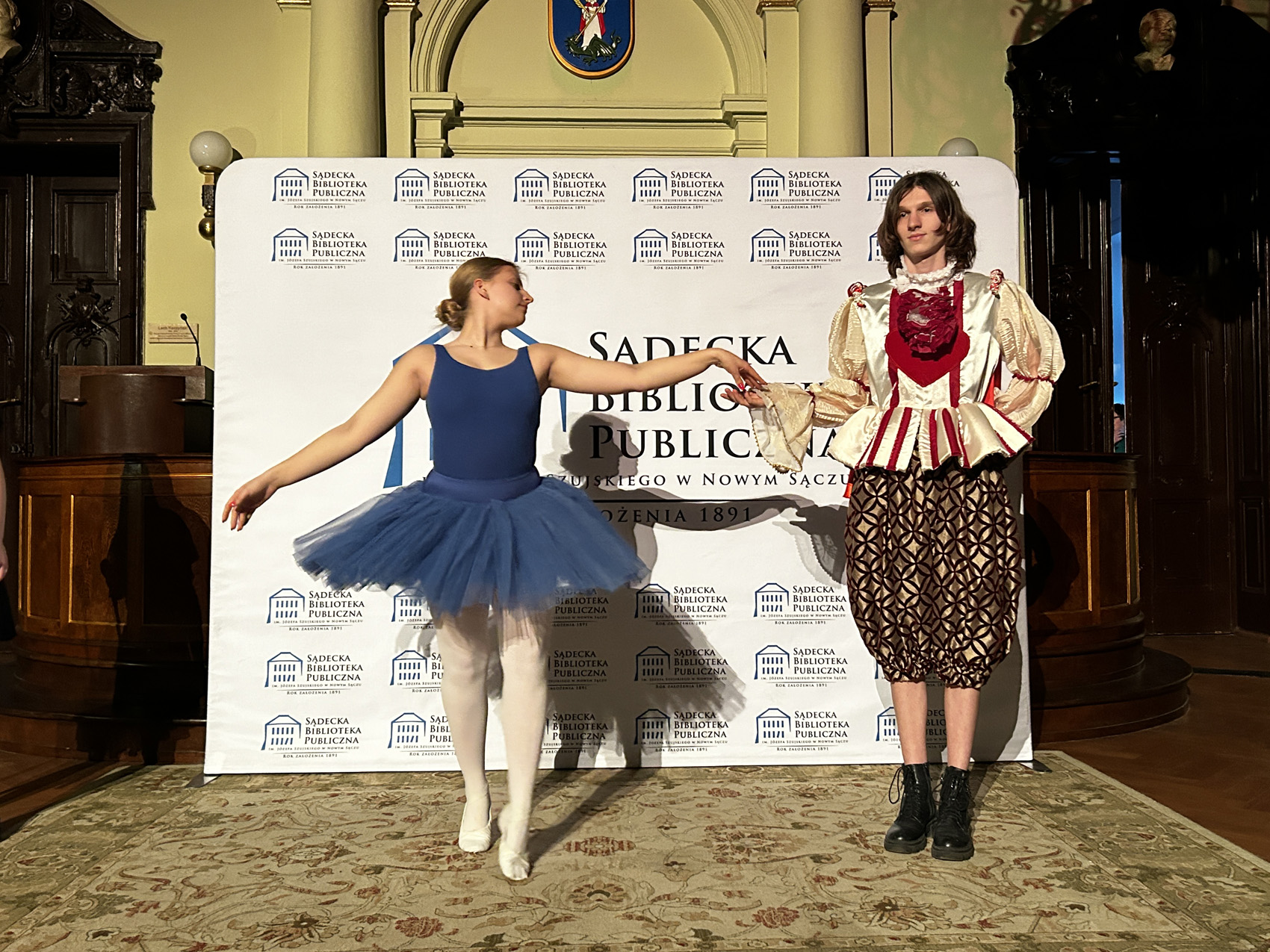 piosenki Jacka Cygana (9) Kobieta i mężczyzna tańczą. Po lewej stronie kobieta w niebiskim stroju baletnicy podaje rękę mężczyźnie stojącemu po prawej stronie. Mężczyzna ma na sobie strój składający się z bufiastej, kremowej bluzki z czerwonym żabotem oraz baskinią oraz brązowych spodni we wzory.