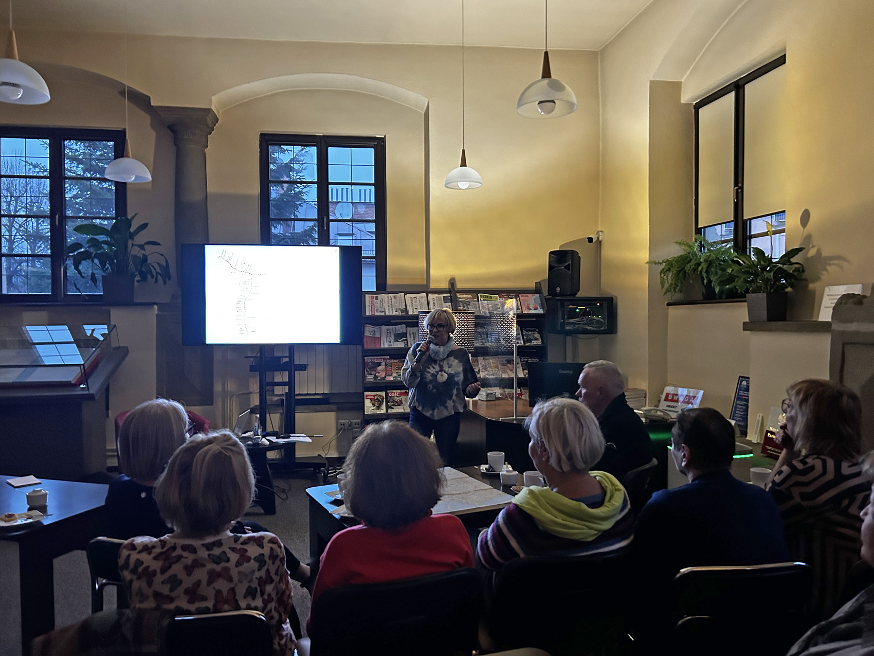 teresa bugaj – podróże na emeryturze (3) Półmrok. Czytelnia w bibliotece. Na krzesłach przy stolikach siedzi grupa 7 osób pokazanych od tyłu. Przed nimi na środku stoi kobieta. Kobieta trzyma w ręce mikrofon blisko ust, mówi do niego. Za kobietą po lewej stronie stoi duży ekran. Po prawej stronie ekranu stoi duży regał z czasopismami.