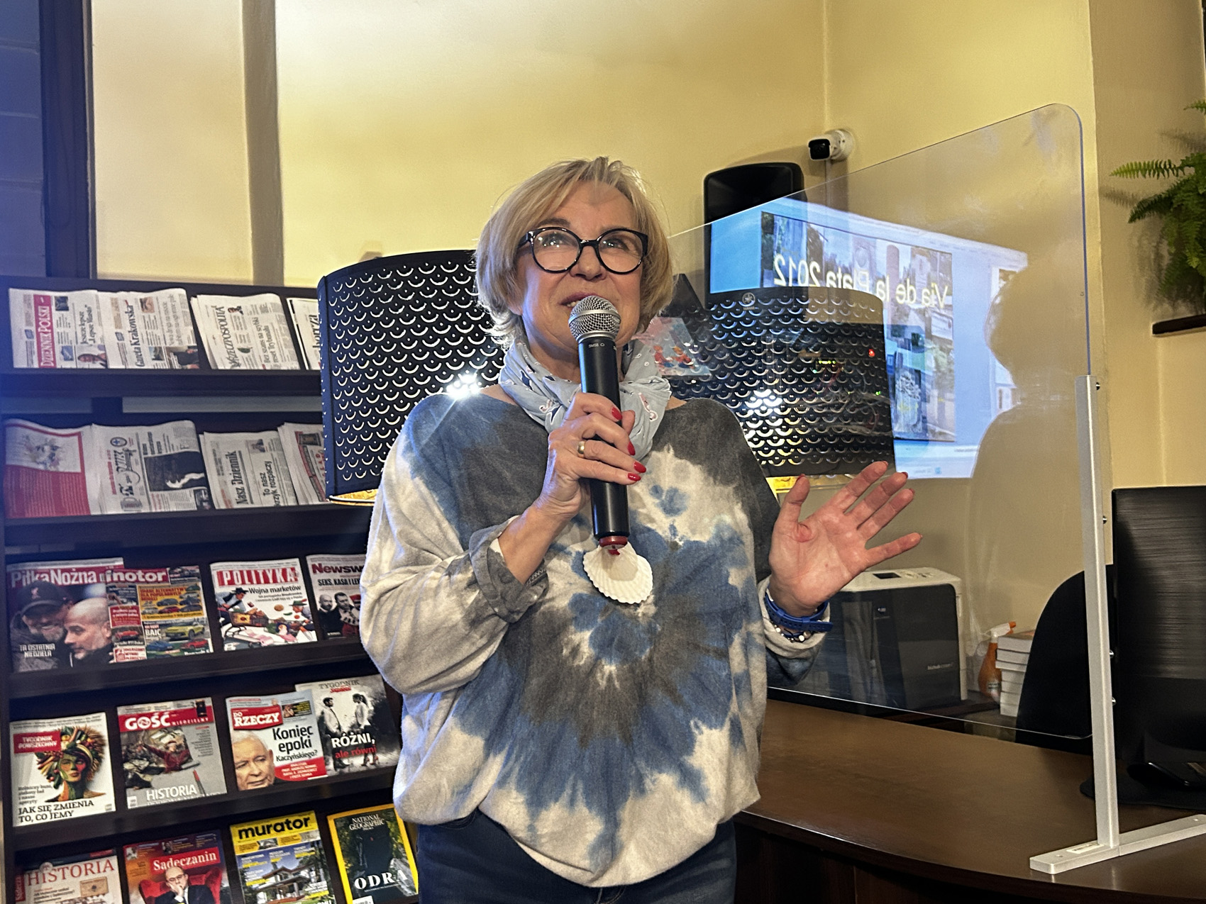 teresa bugaj – podróże na emeryturze (6) W pomieszczeniu znajduje się kobieta pokazana od pasa w górę. Kobieta w jednej ręce trzyma mikrofon blisko ust, mówi do niego. Spogląda w górę. Za kobietą w tle stoi regał z czasopismami.