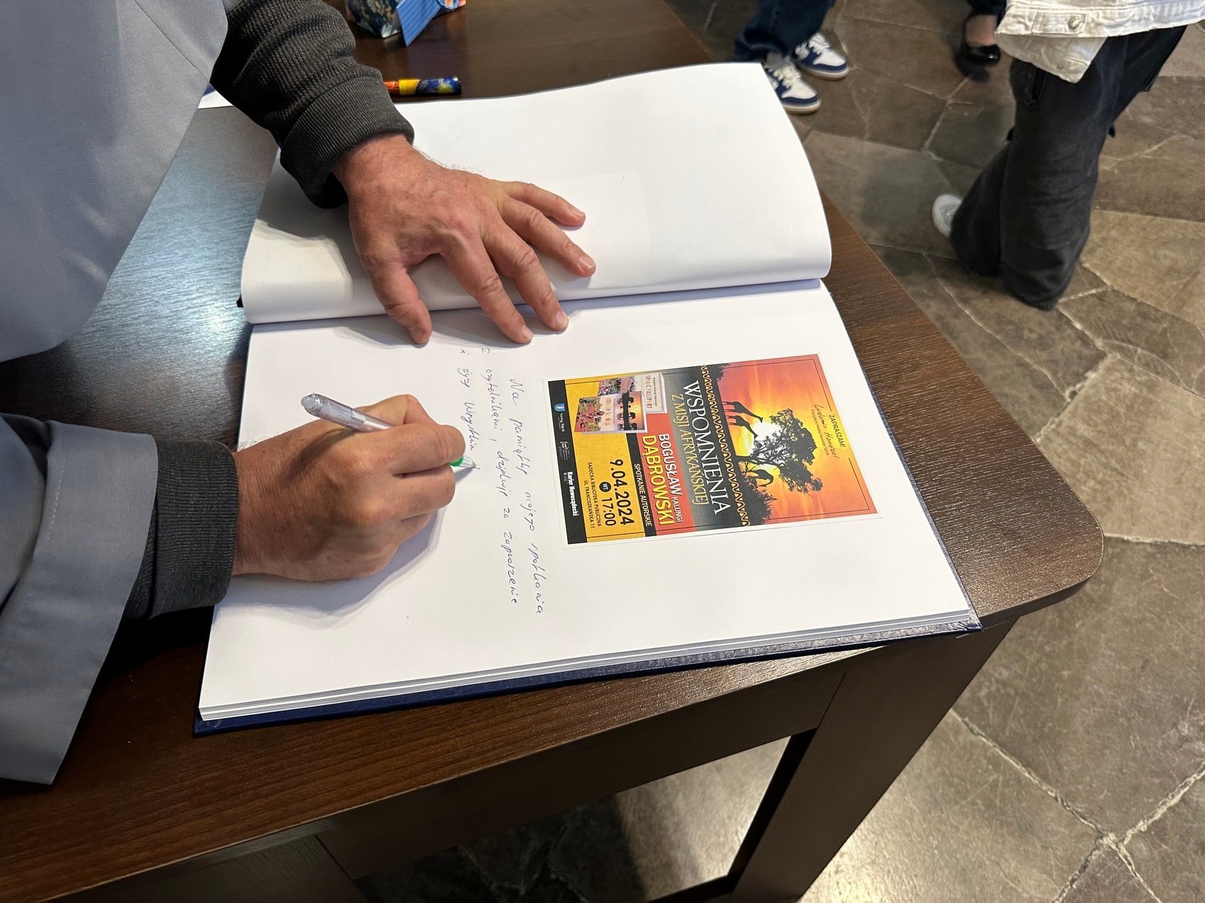 10 Na zdjęciu ukazane są ręce oraz duża księga z wklejonym zdjęciem afrykańskiego krajobrazu. Jedna trzyma długopis i długopis i pisze.