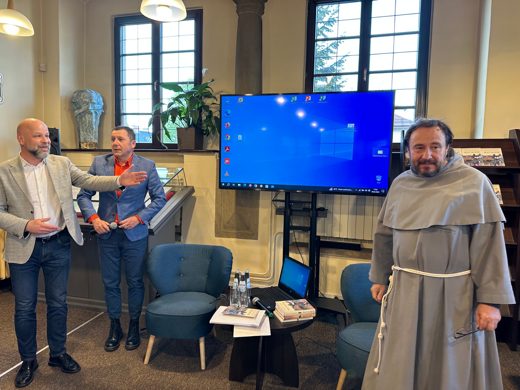 2 Na zdjęciu widać trzech mężczyzn stojących obok siebie. Koło nich ustawiony jest stolik oraz dwa fotele, ekran telewizora z niebieskim tłem oraz regał z książkami.