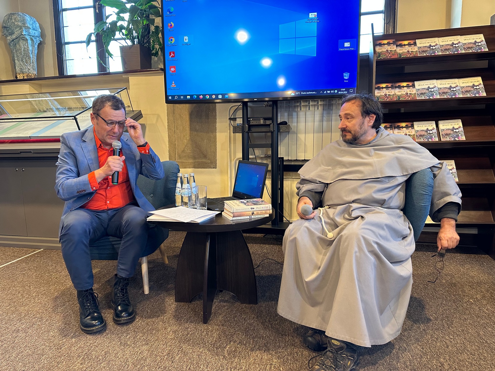 3 Dwóch mężczyzn siedzi patrzy stoliku. Mężczyzna po lewej ubrany w niebieskie spodnie, marynarkę oraz pomarańczową koszulę. Po prawej stronie siedzi mężczyzna w szarym habicie. Między nimi znajduje się ciemny stolik, na którym leżą książki, laptop, szklanki, napoje oraz plik kartek. W tle duży ekran z niebieskim tłem oraz regał z książkami.