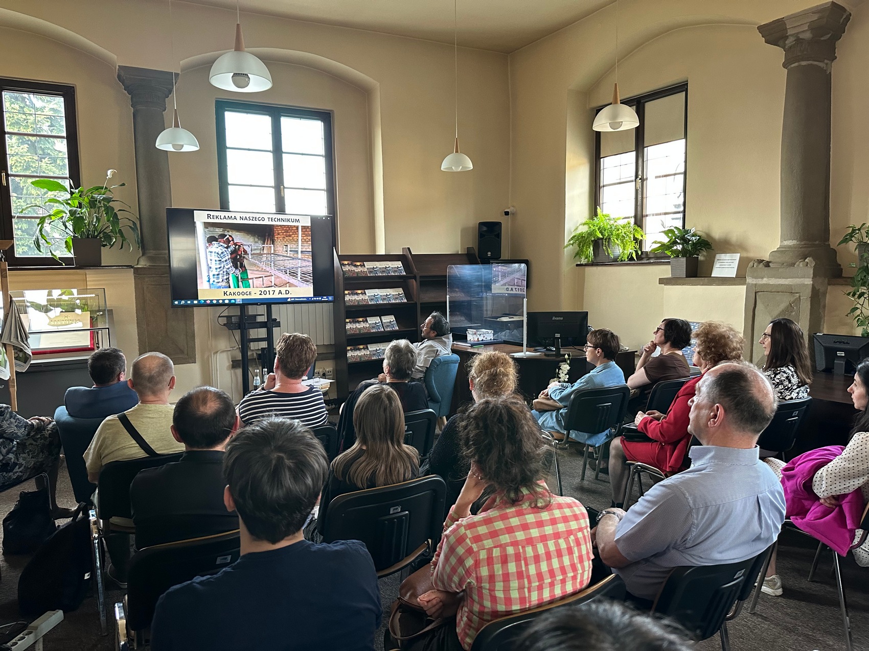 4 Duża sala wypełniona ludźmi siedzącymi na krzesłach. Na przodzie sali ekran telewizor, na którym leci film, dookoła sali okna oraz kwiaty.