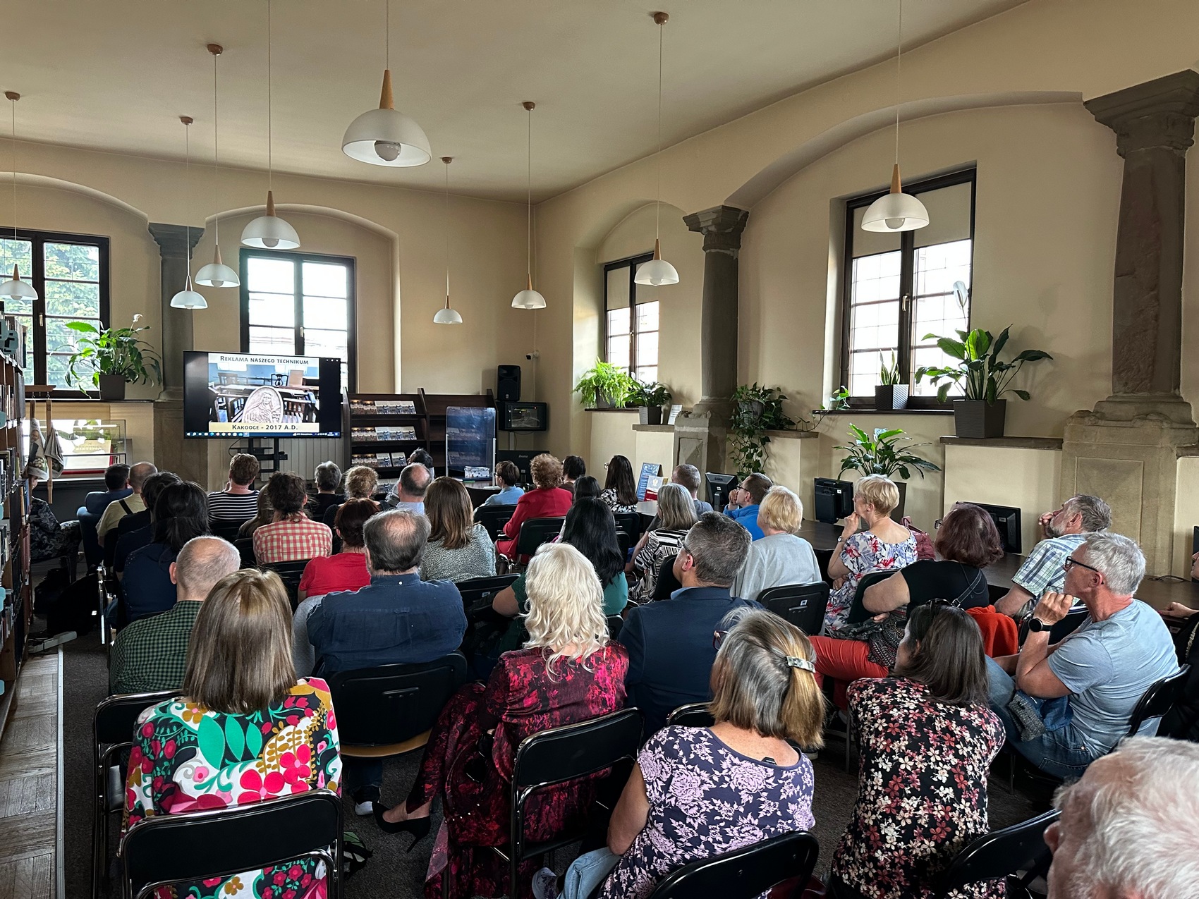 5 Duża sala wypełniona ludźmi siedzącymi na krzesłach. Na przodzie sali ekran telewizor, na którym leci film, dookoła sali okna oraz kwiaty.