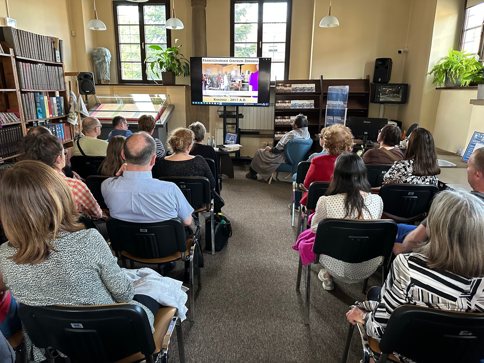 6 Sala pełna ludzi siedzących w dwóch rzędach. Na przodzie siedzi w fotelu mężczyzna w habicie odwrócony tyłem, patrzący na ekran telewizora.