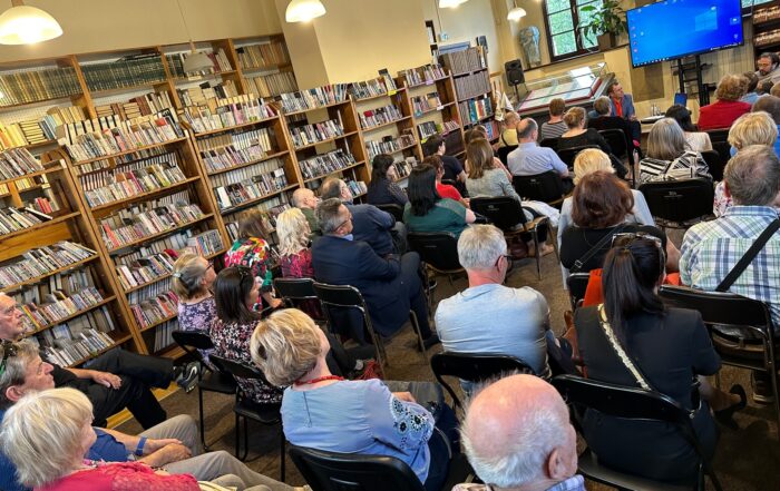 Sala pełna ludzi siedzących na krzesłach. Po lewej stronie ustawione regały z książkami. W oddali dwóch mężczyzn siedzących na fotelach, za nimi ekran z niebieskim tłem.