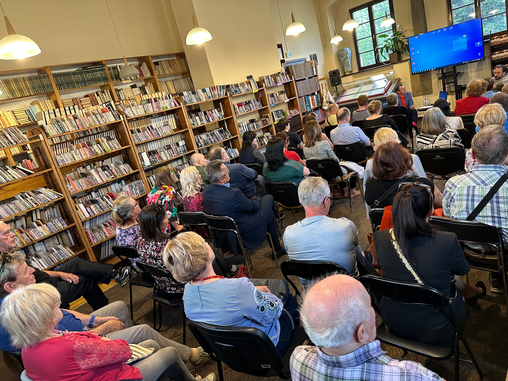 Sala pełna ludzi siedzących na krzesłach. Po lewej stronie ustawione regały z książkami. W oddali dwóch mężczyzn siedzących na fotelach, za nimi ekran z niebieskim tłem.