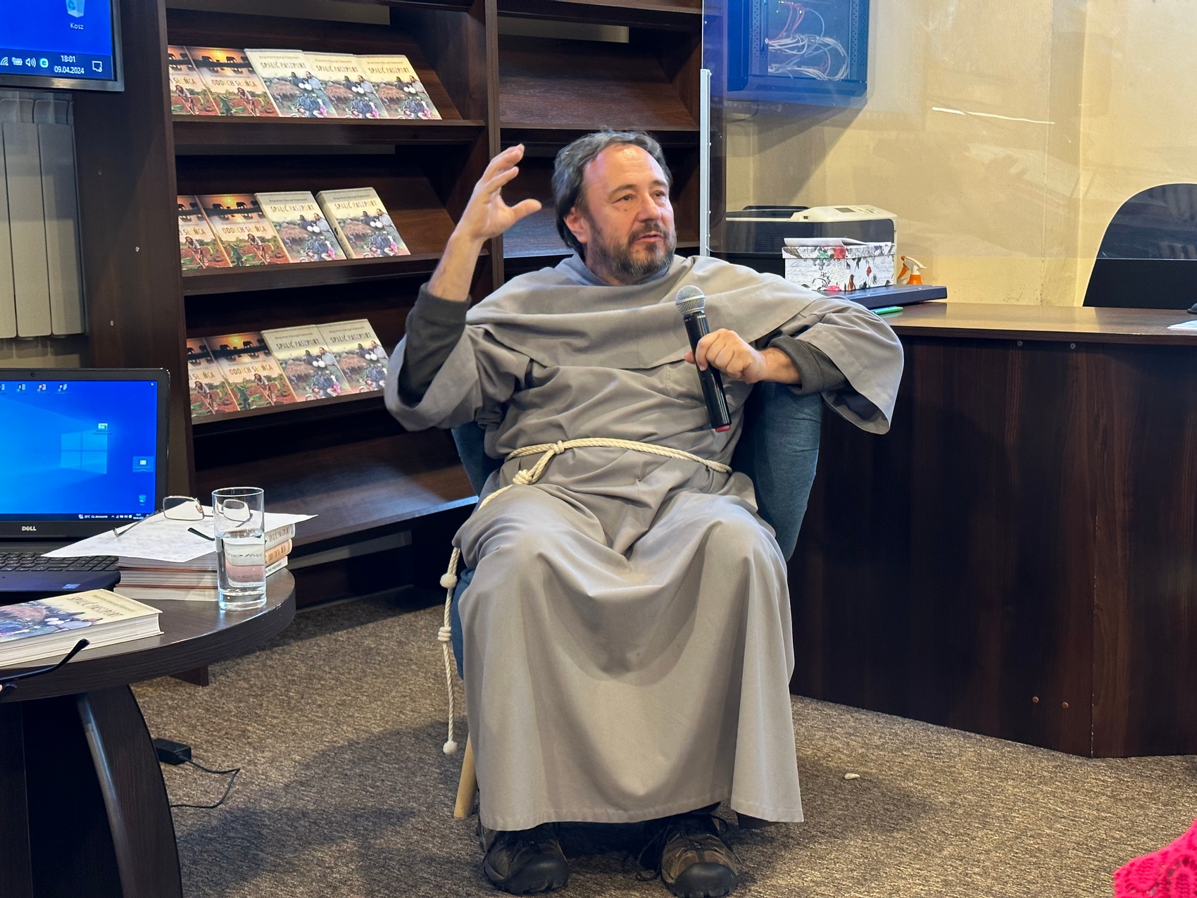 8 Mężczyzna w szarym habicie siedzi na fotelu i mówi do mikrofonu. Po lewej stronie widoczny jest fragment stolika, na którym stoją książki, laptop, szklanka oraz okulary. W tle regał z książkami oraz biurko.