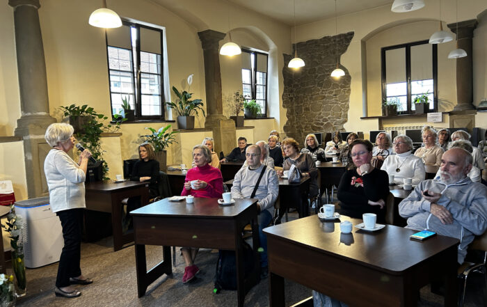 Sala czytelni biblioteki, po lewej stronie stoi kobieta w białym swetrze, mówi do mikrofonu. Przed nią przy stołach siedzą ludzie, słuchają i popijają napoje.
