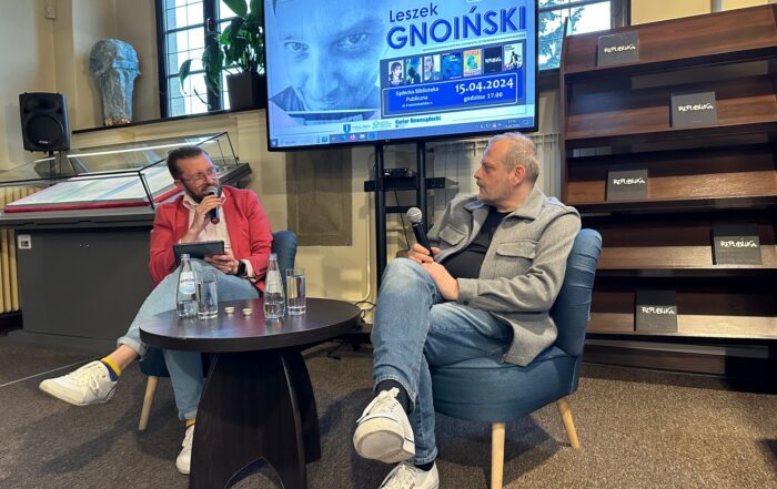 Dwóch mężczyzn rozmawia ze sobą siedząc przy stoliku, obydwoje trzymają mikrofony. Obok ustawione są regały z książkami oraz ekran z wizerunkiem jednego z mężczyzn.