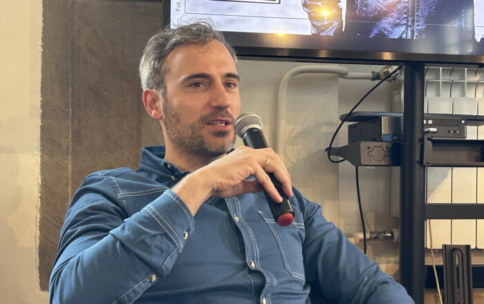 Siedzący mężczyzna widoczny od pasa w górę, w ręku trzyma mikrofon.