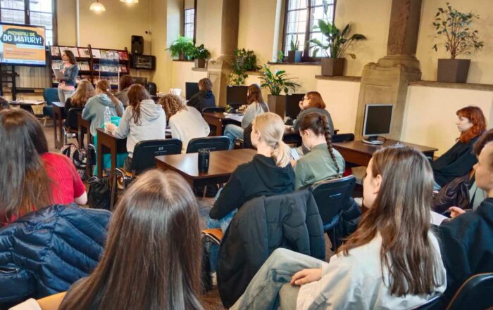 Sala wypożyczalni sądeckiej biblioteki, grupa zgromadzonych ludzi siedzi przy biurkach, słuchając kobiety stojącej przed nimi.