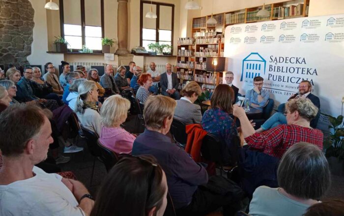 Duża grupa ludzi siedzących słucha trzech mężczyzn siedzących przed nimi. W tle widoczne są książki oraz audiobooki oraz logo Sądeckiej Biblioteki Publicznej.