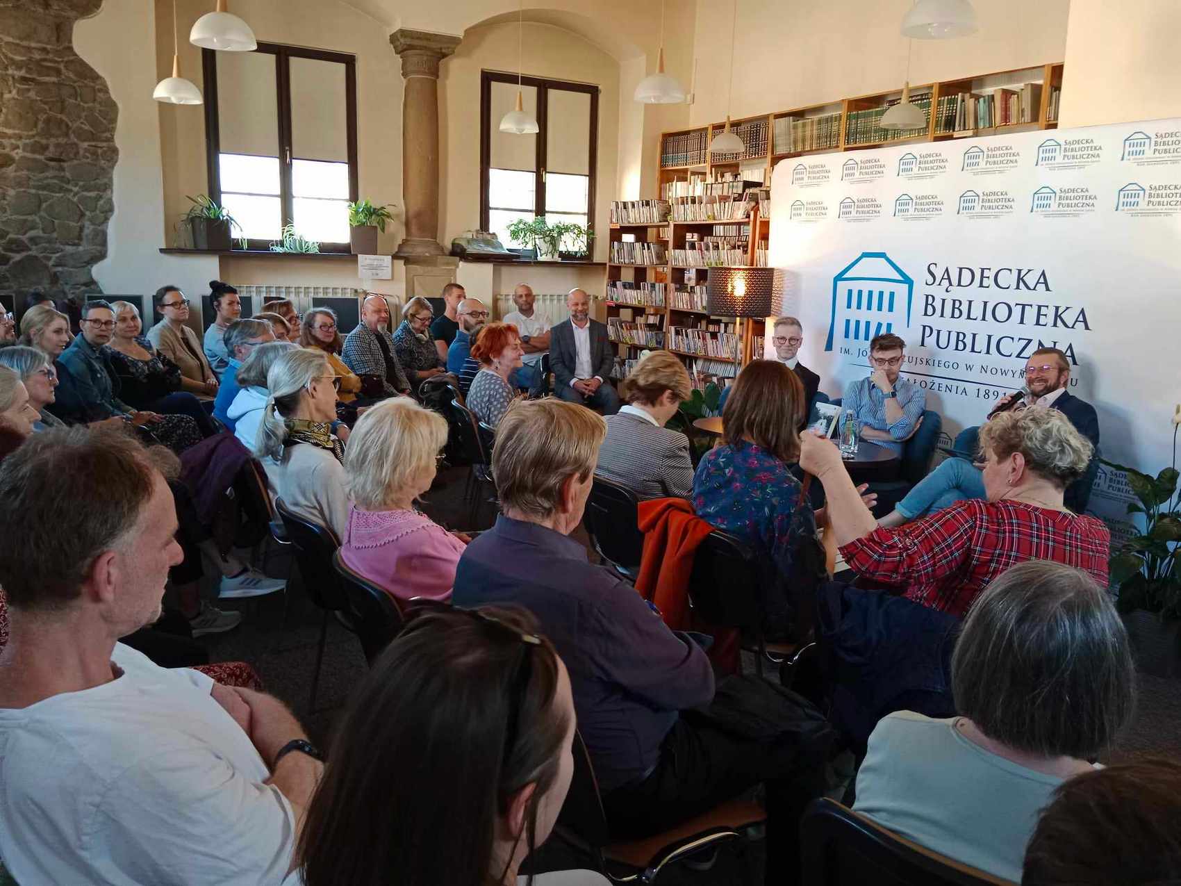 Duża grupa ludzi siedzących słucha trzech mężczyzn siedzących przed nimi. W tle widoczne są książki oraz audiobooki oraz logo Sądeckiej Biblioteki Publicznej.