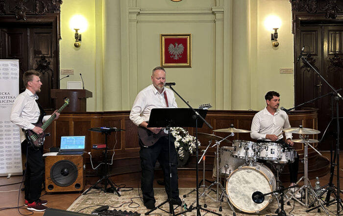 Trzech mężczyzn grających na instrumentach. Mężczyzna po prawej stronie siedzi na krześle i gra na perkusji. Mężczyzna po środku gra na gitarze i śpiewa do mikrofonu. Mężczyzna po lewej stronie również gra na gitarze.