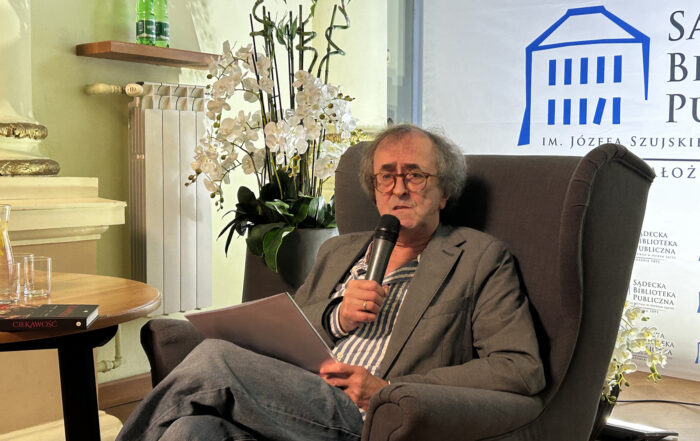 Mężczyzna siedzi na szarym fotelu przy stoliku, w ręce trzyma mikrofon, za nim widnieje fragment loga Sądeckiej Biblioteki Publicznej.