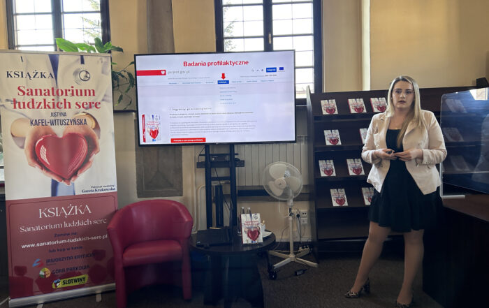 Sala z dużymi oknami. Po prawej stronie stoi kobieta w blond włosach. Za nią na regale umieszczone są książki z sercem na okładce. Obok widnieje telewizor z prezentacją na temat badań profilaktycznych. Z boku telewizora rozłożony jest plakat, na którym postać w białym kitlu lekarskim trzyma czerwone serce. oraz napis: Sanatorium ludzkich serc, Justyna Kafel-Wituszyńska.