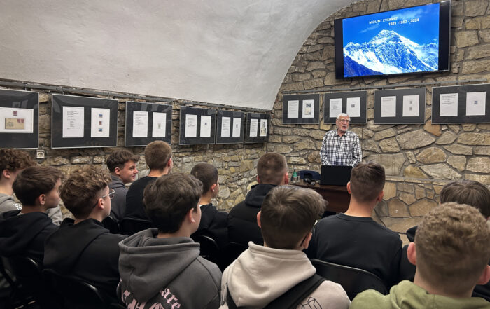 Grupa ludzi siedzących tyłem patrzy na mężczyznę stojącego przed nimi. Na ścianach wiszą metalowe prowadnice z ramami, w których widnieją znaczki. Wyżej na ścianie zamontowany jest telewizor, na którym widnieją ośnieżone szczyty gór oraz napis: Mount Everest 1921 - 1953 - 2024