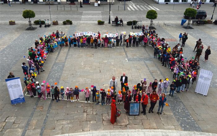 Zdjęcie z lotu ptaka. Na placu miejskim stoją dzieci tworząc kształt książki. Część dzieci trzyma transparent z napisem "Biblioteka - miejsce na czasie". Po bokach stoją dwa banery z logotypem Sądeckiej Biblioteki Publicznej. Przed dziećmi stoi postać w czerwonej pelerynie, obok niej kilka osób z czerwonymi elementami stroju trzyma duży plakat.