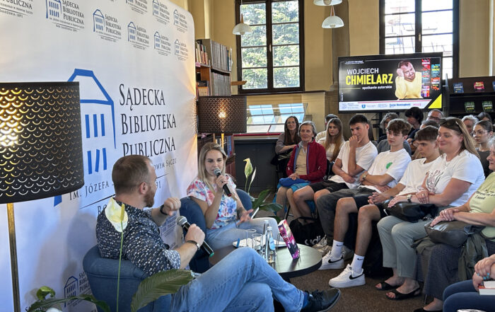 Grupa ludzi przygląda się mężczyźnie oraz kobiecie siedzących przed nimi na krzesłach przy stoliku. W tyle widać telewizor z mężczyznom w czerwonym swetrze, obok napis: Wojciech Chmielarz, spotkanie autorskie.