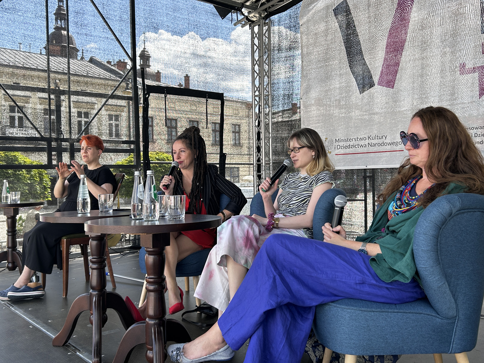 Trzy kobiety siedzą na scenie przy stoliku, wszystkie trzymają w rękach mikrofony. Obok po lewej stronie siedzi kobieta tłumacząca rozmowę na język migowy.