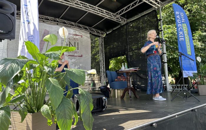 Na scenie stoi kobieta w niebieskiej sukience mówiąca do mikrofonu. Za nią ustawiony jest stolik oraz niebieski fotel. Po lewej stronie ustawiony jest kwiat w doniczce za którym siedzi kobieta w granatowej sukience.