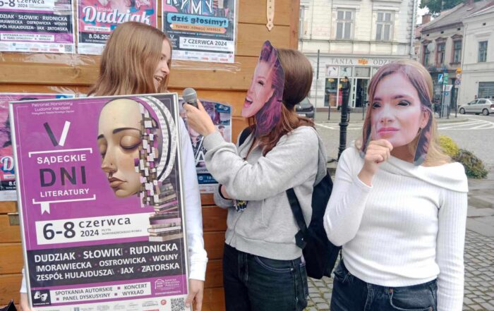 Grupa trzech osób stojących na chodniku. Dwie kobiety stojące po prawej stronie mają założone maski na twarzach. Kobieta stojąca w środku trzyma w ręku mikrofon, zwraca się w stronę kobiety stojącej po lewej stronie. Ta z uśmiechem patrzy na nią, trzymając ramę z plakatem w niej umieszczonym. W tle fragment drewnianego domku oraz zabytkowych kamienic.