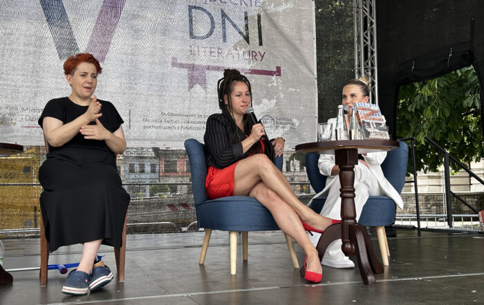 Na scenie siedzą trzy kobiety. Kobieta po prawej stronie w białej marynarce, częściowo zasłonięta przez stojący stolik, mówi do mikrofonu, kobieta po lewej stronie, patrzy w dół, trzymając mikrofon blisko ust. Na stoliku ustawiona jest książka oraz napoje. Po lewej stronie siedzi kobieta, tłumacząca rozmowę na język migowy.