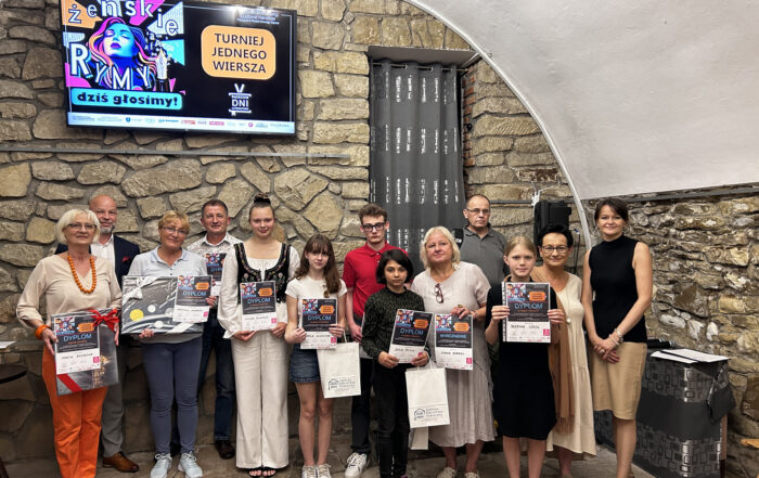 Grupa ludzi stoi pod ściana, trzymając w rękach dyplomy. Nad nimi na kamiennej ścianie zawieszony jest telewizor na którym widnieje podobizna kobiety oraz napis: Żeńskie rymy dziś głosimy. Turniej jednego wiersza.