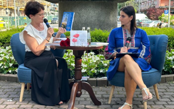 Przy stoliku, na niebieskich fotelach siedzą dwie kobiety. Kobieta po prawej stronie w niebieskiej sukience, patrzy na kobietę w białej bluzce siedzącą obok, trzymającą w jednej ręce mikrofon, w drugiej książkę. Za nimi fragment pomnika otoczonego kwiatami.