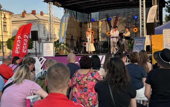 Zgromadzona publiczność stoi oraz siedzi pod oświetloną sceną, na której stoi grupa ludzi w towarzystwie sprzętów muzycznych. Po lewej stronie widoczne są zabytkowe kamienice miasta.