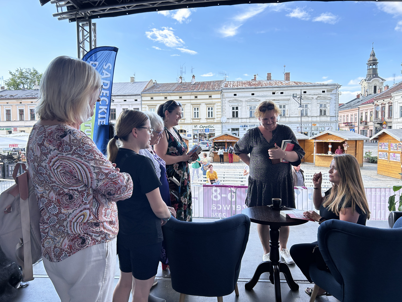 Olga Rudnicka (1) Na scenie w centrum miasta siedzi kobieta przy stoliku, przed nią leży książka, wokół zgromadzone kobiety przysłuchują się. W tle widoczne są zabytkowe kamienice.