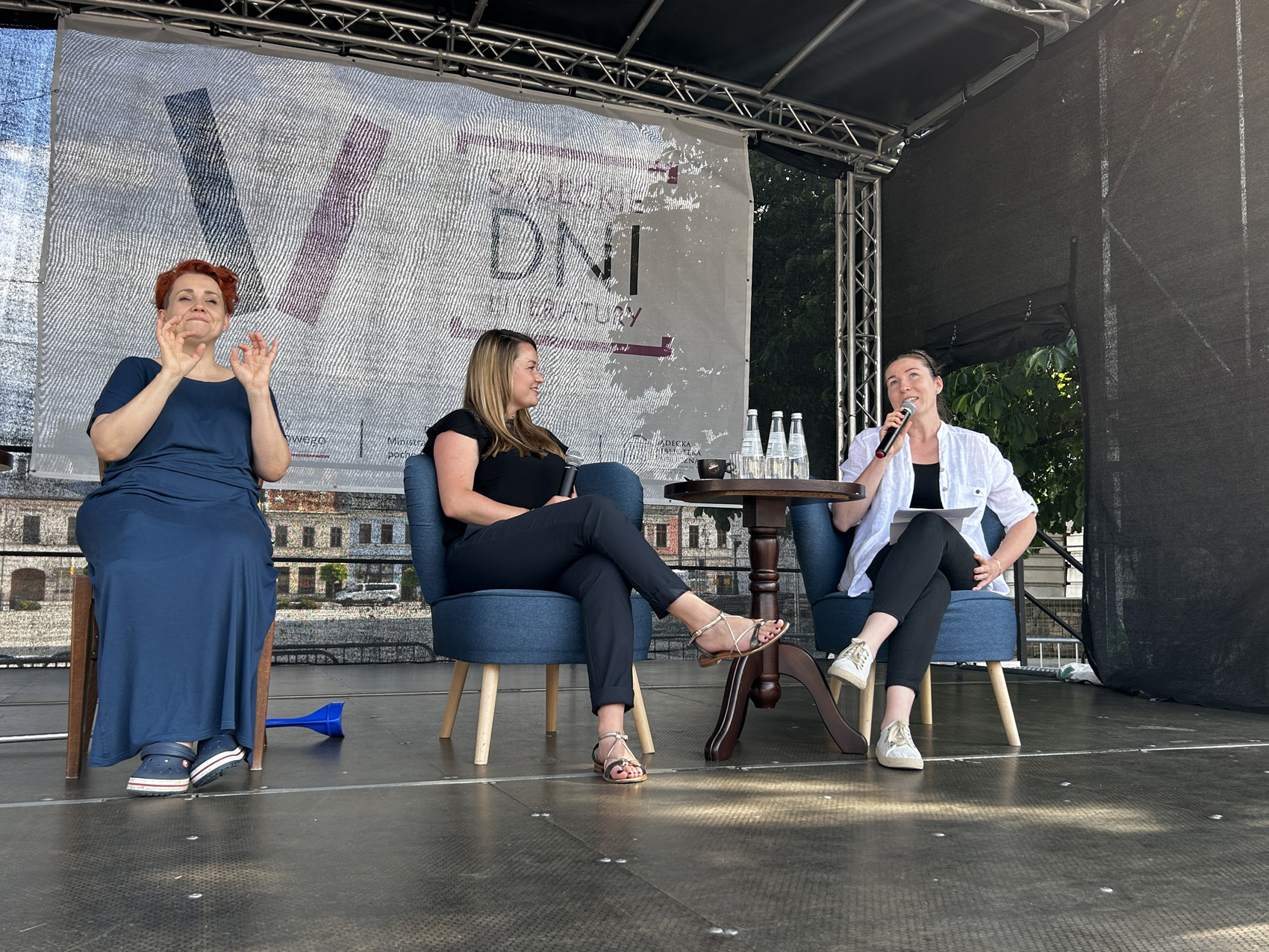 Olga Rudnicka (2) Na scenie siedzą trzy kobiety. Po prawej stronie umieszczony jest stolik, przy którym siedzą dwie kobiety, jedna z nich mówi do mikrofonu. po lewej stronie kobieta tłumaczy rozmowę na język migowy.