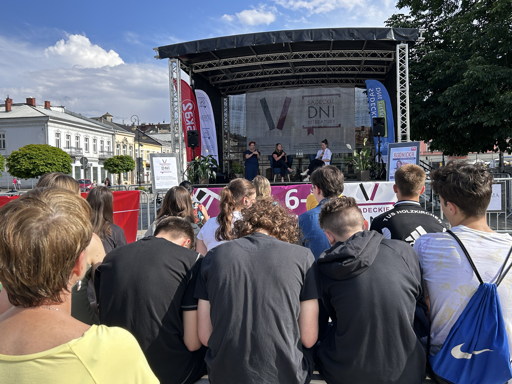 Olga Rudnicka (3) Zwrócona tyłem publiczność patrzy w stronę sceny, na której siedzą trzy kobiety. Po prawej stronie kobieta w białej koszuli patrzy w dół, siedząca obok niej przy stoliku kobieta mówi do mikrofonu. Z lewej strony sceny siedzi kobieta w granatowej sukience, tłumacząc rozmowę na język migowy. Ze sceny widoczny jest napis: V Sądeckie Dni Literatury. Wokół sceny widoczne są zabytkowe kamienice oraz fragment drzewa.
