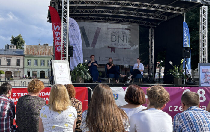 Grupa ludzi licznie zgromadzona siedzi pod sceną. patrzy na kobiety przemawiające ze sceny oraz siedzącą obok niej tłumaczkę migową.