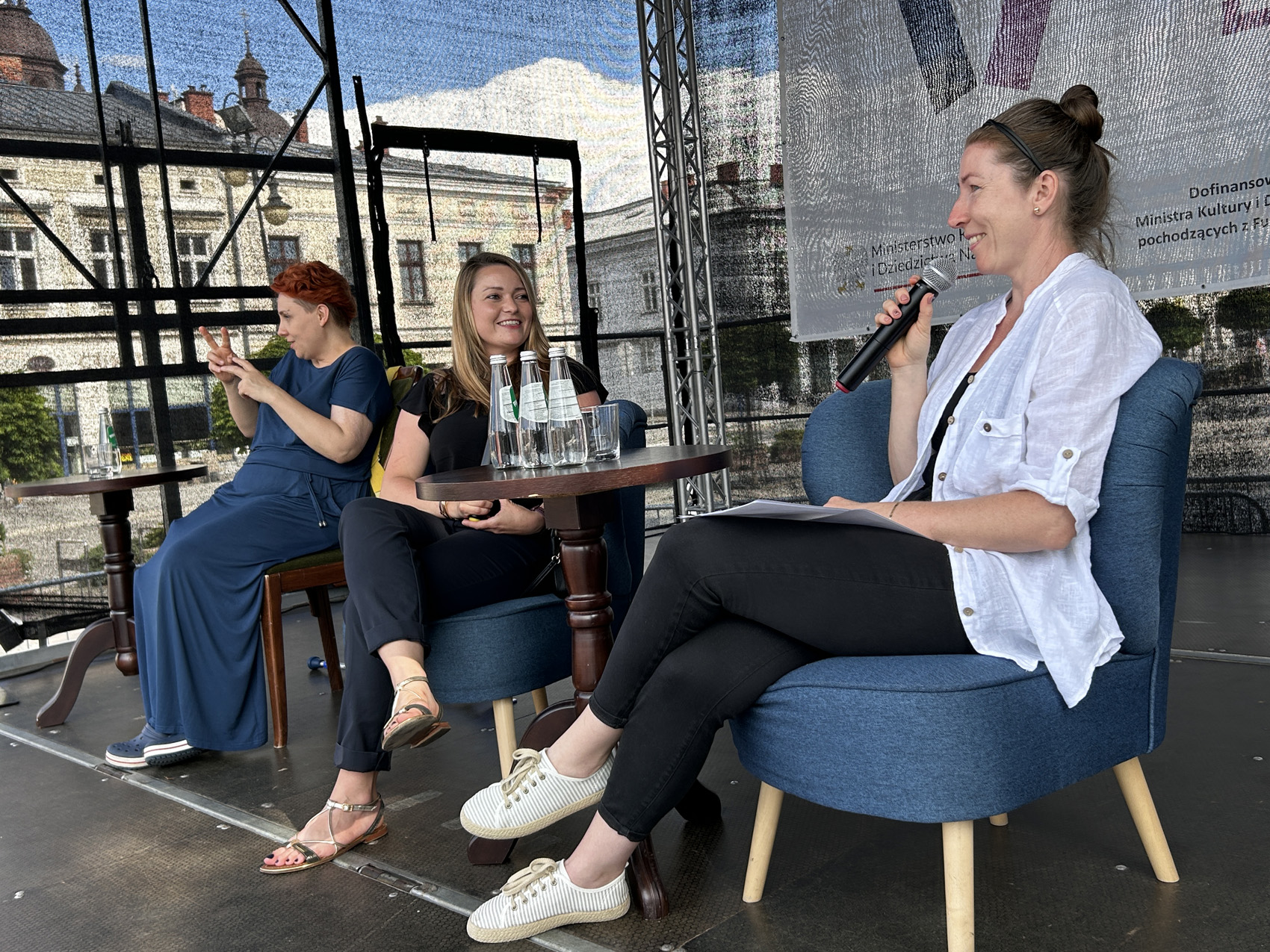 Olga Rudnicka (5) Na scenie siedzą trzy kobiety. Po prawej stronie umieszczony jest stolik, przy którym siedzą dwie kobiety, jedna z nich mówi do mikrofonu. po lewej stronie kobieta tłumaczy rozmowę na język migowy.