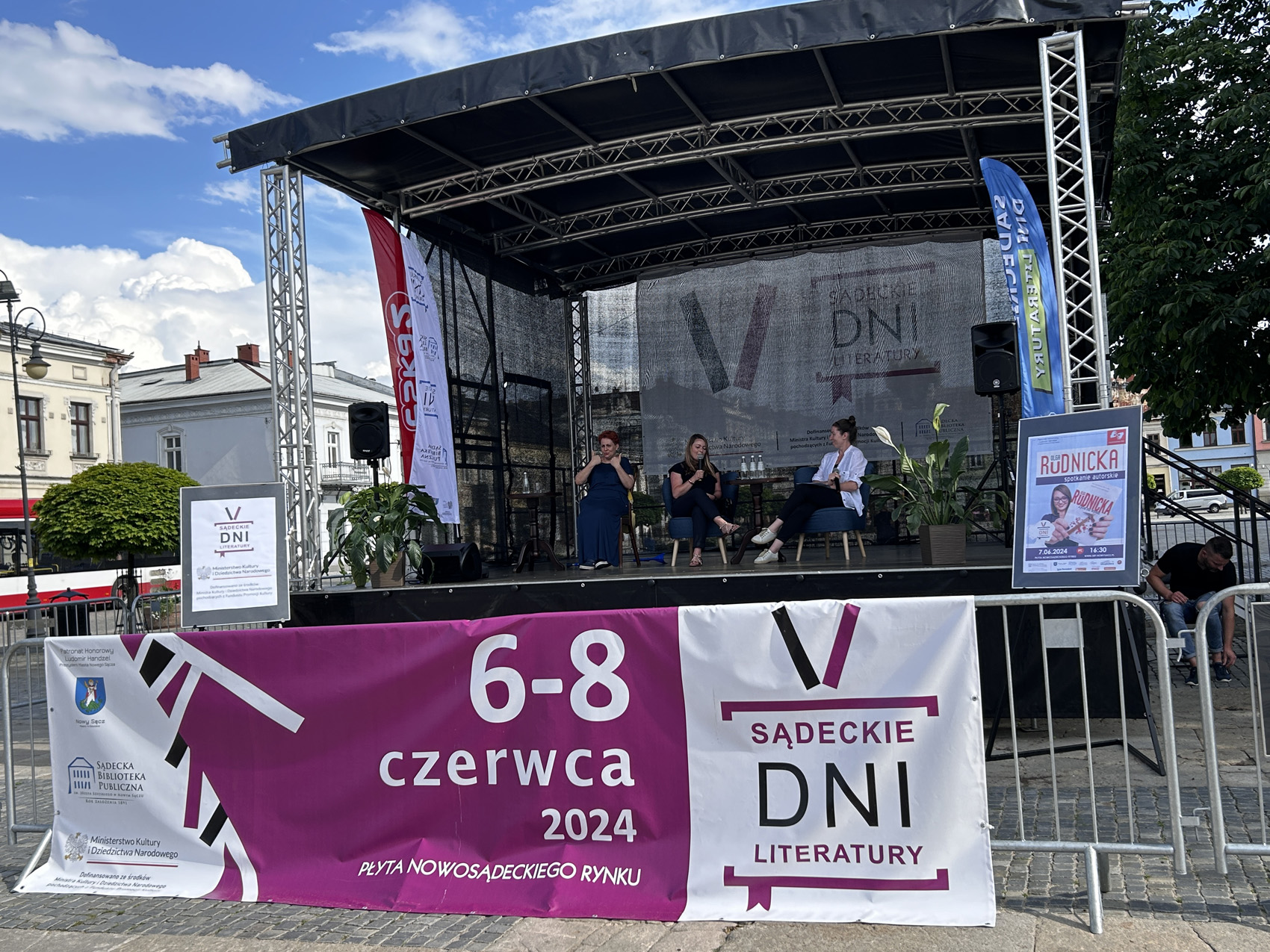 Olga Rudnicka (6) Na scenie siedzą trzy kobiety. Na dole umieszczony jest baner z napisem: 6-8 czerwca 2024, płyta nowosądeckiego rynku, V Sądeckie Dni Literatury.