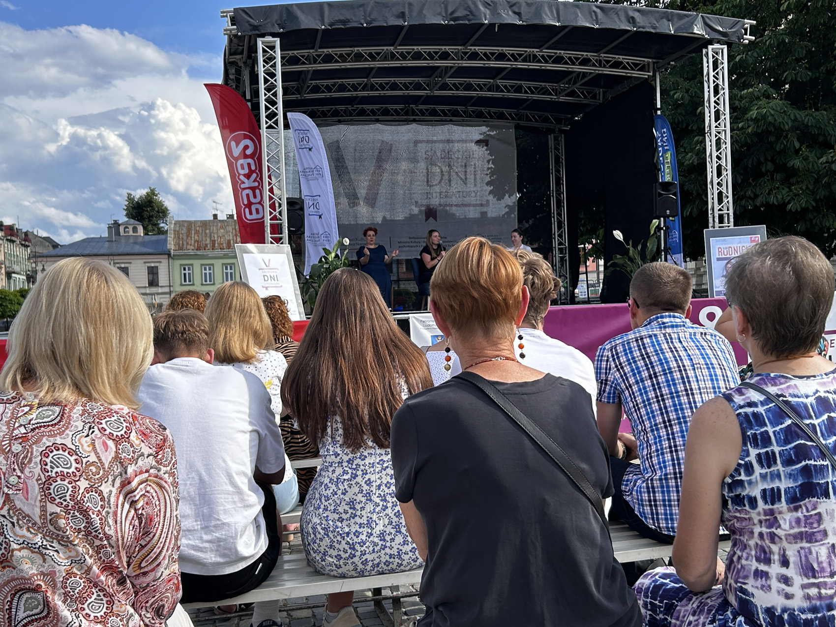 Olga Rudnicka (7) Grupa ludzi zgromadzona pod sceną przysłuchują się dwóm kobietom rozmawiającym na scenie. Obok nich po lewej stronie siedzi kobieta, tłumacząca na język migowy.