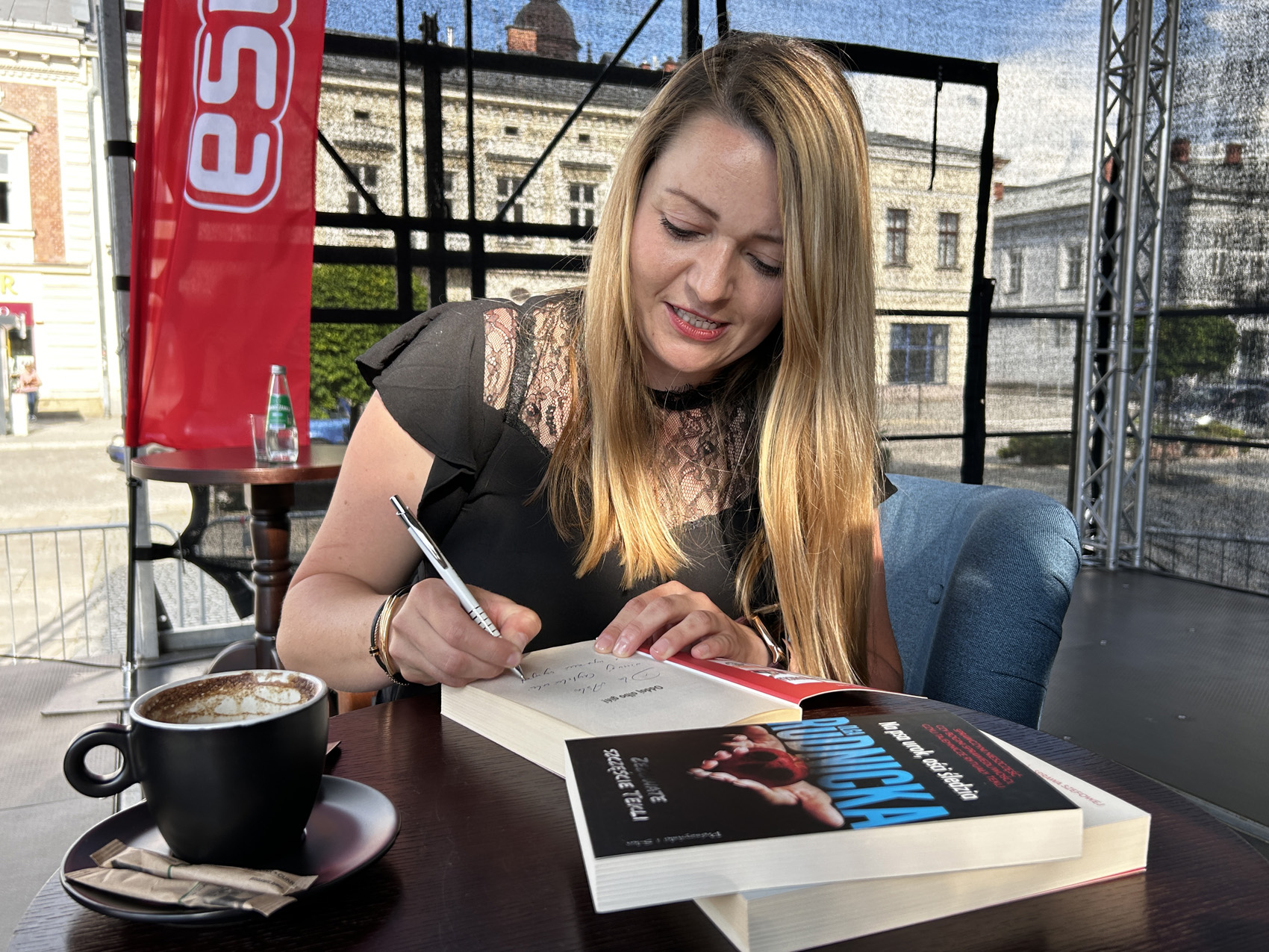 Olga Rudnicka (9) Kobieta w czarnej bluzce siedzi na fotelu, podpisuje umieszczoną na stoliku książkę. Obok widnieje filiżanką z kawą.
